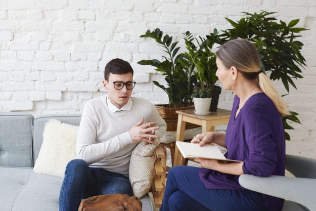 candid-shot-young-man-glasses-talking-about-his-problems-during-psychological-therapy-session-sitting-coach-while-mature-female-psychologist-with-copybook-listening-him-making-notes(1)
